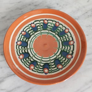 Peut inclure: Une assiette ronde en céramique orange avec un motif géométrique blanc, vert et bleu. Le motif présente un cercle central avec des lignes rayonnantes et des formes de fleurs stylisées.