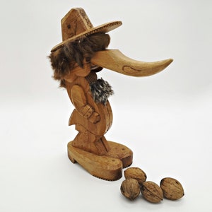 May include: Wooden nutcracker figurine with a long nose, wearing a hat and fur trim. The nutcracker is standing on a base with four walnuts in front of it.