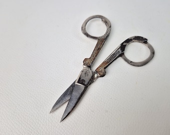 May include: Vintage silver-toned scissors with textured handles and circular finger holes. The blades are sharp and pointed, ideal for detailed cutting. The scissors are likely used for sewing or embroidery.