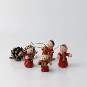 Könnte beinhalten: Eine Sammlung kleiner, handbemalter Weihnachtsornamente aus Holz. Die Ornamente umfassen rot gekleidete Figuren, einen Schneemann und einen Tannenzapfen. Jedes Ornament hat eine goldene Schnur zum Aufhängen. Die Figuren haben bemalte Gesichter und goldene Akzente.