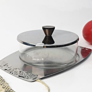 Könnte beinhalten: Eine silberne Butterdose mit einem Glasdeckel und einem Holzgriff. Die Dose steht auf einem silbernen Tablett mit einem dekorativen Rand.