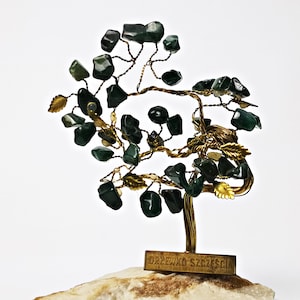Könnte beinhalten: Eine Draht-Baumskulptur mit grünen Steinen, goldenen Blättern und einem Sockel aus Naturstein. Der Sockel trägt eine kleine Plakette mit der Aufschrift "Drzewko Szczęścia".