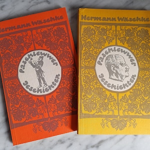Könnte beinhalten: Zwei antike gebundene Bücher mit orangefarbenen und gelben Einbänden. Die Einbände zeigen kunstvolle florale Muster und den Titel "Paschlewwer Geschichten" in einem Kreis mit einem Porträt eines Mannes. Der Name des Autors, "Hermann Wissekke", ist oben auf jedem Einband gedruckt.