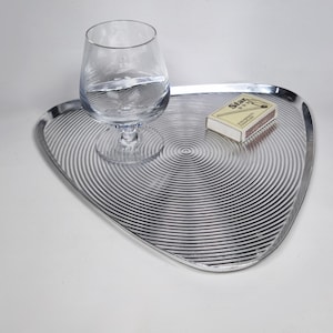 Könnte beinhalten: Ein dreieckiges, silberfarbenes Metalltablett mit einem konzentrischen, kreisförmigen Rillenmuster. Ein klares Glasschwenker steht auf der linken Seite des Tabletts, und eine rechteckige weiße Streichholzschachtel mit dem Aufdruck Star liegt rechts.
