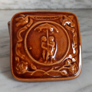 May include: A square, brown ceramic tile with a glossy finish. The tile features a central circular design of two figures under an umbrella, surrounded by floral and ribbon motifs. The word "CIECHOCINEK" is at the top.