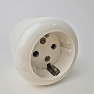 May include: A white electrical outlet with three circular holes and a grounding pin. The outlet is made of a glossy, rounded material and is set against a plain white background. The outlet is designed to receive a plug.
