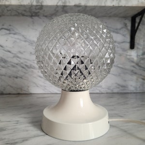 May include: A white table lamp with a clear glass globe shade featuring a diamond pattern. The base is white and round.