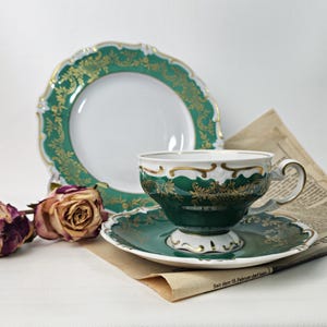 Könnte beinhalten: Ein Vintage-Teeservice mit grünem und goldenem Design. Das Set umfasst eine Tasse, eine Untertasse und einen Teller. Die Tasse und Untertasse liegen auf einer Zeitung. Getrocknete Rosen sind im Vordergrund.