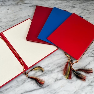 May include: An open red presentation folder with cream pages, next to three closed folders in red and blue. Each folder has a decorative tassel with black, yellow, and red threads. The folders are on a marble surface.