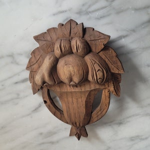 May include: A carved wooden wall hanging depicting a basket overflowing with fruit. The fruit includes pears and round fruits, all carved in a detailed, realistic style. The piece is made of a light brown wood and has a rustic, vintage aesthetic.