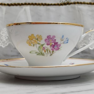 Può includere: Una tazza da tè e un piattino bianchi con un bordo dorato. La tazza da tè ha un motivo floreale con fiori gialli, rosa e blu.