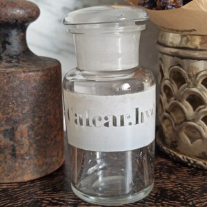 May include: A clear glass bottle with a white label that says "Caleaur: Ivy". The bottle is sitting on a wooden surface next to a rusty metal weight.