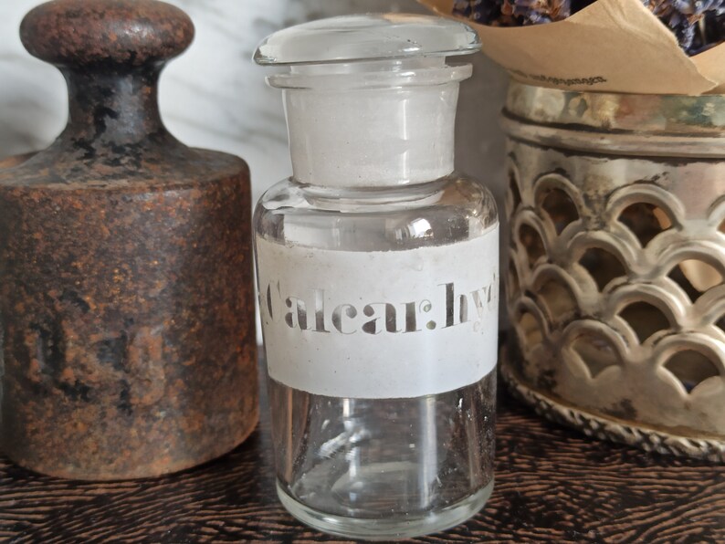 May include: A clear glass bottle with a white label that says "Caleaur: Ivy". The bottle is sitting on a wooden surface next to a rusty metal weight.