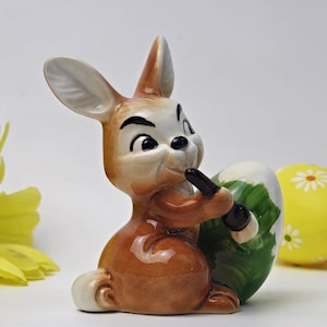 Könnte beinhalten: Eine Keramik-Osterhasenfigur, die ein grünes und weißes Osterei hält. Der Hase hat braunes Fell und einen schelmischen Ausdruck.