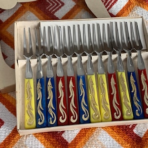 May include: A boxed set of vintage forks. The forks have stainless steel tines and handles in alternating colours of yellow, blue, and red, with a gold decorative design. The box is beige.