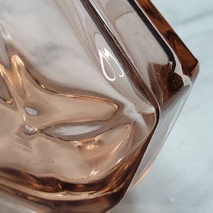 May include: A close-up of a clear glass vase with a textured surface. The vase is a light pink colour and has a square shape.