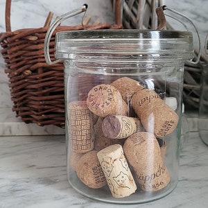 May include: A glass jar with a metal lid filled with a collection of used wine corks. The corks are various sizes and have different markings and text, including "Dargetz" and "Kappelbrunner".
