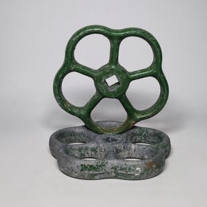 Può includere: Due volanti per valvole industriali con cinque razze arrotondate e un foro centrale quadrato. Una ruota è verniciata in verde scuro lucido, mentre l'altra è in metallo grigio strutturato e usurato con residui di vernice verde.