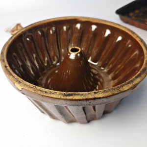 May include: A brown ceramic bundt cake tin with a central cone and ridged sides. The tin has a dark brown glaze and a lighter rim. A baking tray is visible in the background.