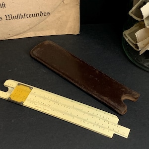 Może przedstawiać: Antyczna suwmiarka z żółtą i kremową skalą, obok ciemnobrązowego skórzanego etui. Suwmiarka posiada szczegółowe oznaczenia i metalową ramę. Przedmiot kolekcjonerski.