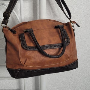 May include: A brown leather satchel handbag with a black trim and a long strap. The bag has a top zipper closure and two top handles.