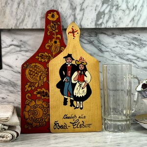 May include: Two wooden cutting boards with painted folk art designs. One is red with gold floral patterns, the other is light wood with a couple in traditional clothing. Text reads "Gruss aus Bad-Elster".
