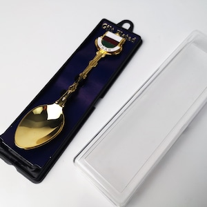 以下が含まれることがあります： 青と白の旗のデザインと「OMAN」の文字が書かれた金色の記念スプーン。スプーンは、「Gift Tried」と書かれた黒いベルベットの箱に入っています。スプーンは透明なプラスチックの箱の隣にあります。