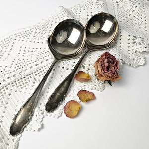 May include: Two silver serving spoons on a white crocheted doily with a dried rose and petals.