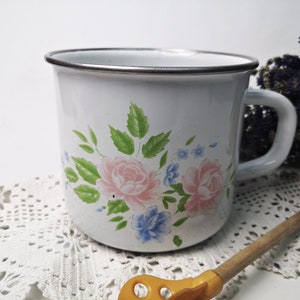 Könnte beinhalten: Weiße Emailtasse mit einem Blumendesign mit rosa Rosen, grünen Blättern und blauen Blüten. Die Tasse hat einen silbernen Rand und einen Henkel.