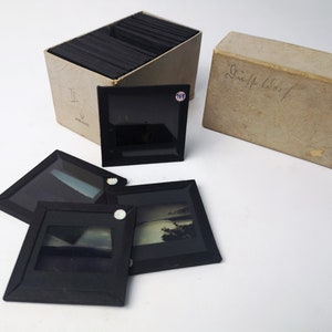 May include: A box of vintage glass slides with a handwritten label that says "Diapositives". The box is open and four slides are lying on a white surface. The slides are black with a clear glass insert.