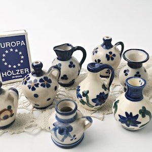 Könnte beinhalten: Eine Sammlung von Miniatur-Keramikkrügen und Vasen in blau-weißen Blumenmustern. Die Krüge haben einen Henkel und einen Ausguss, während die Vasen rund sind und eine schmale Öffnung haben. Die Krüge und Vasen sind auf einer weißen Spitzentischdecke angeordnet.