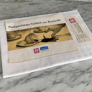 Könnte beinhalten: Eine weiße Tischdecke aus Baumwolle mit Rautenmuster, begleitet von einer Vintage-Werbung und einem Etikett. Die Werbung zeigt eine Zeichnung einer verschütteten Kaffeetasse. Der Text auf der Werbung lautet "Fleckgeschütztes Tischtuch aus Baumwolle".