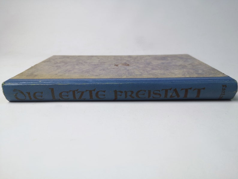 May include: A blue hardcover book with the title "Die letzte Freistatt" printed in gold lettering on the spine.
