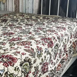 Peut inclure: Une couverture blanche avec un motif floral dans les tons de rouge, vert et marron. La couverture a une bordure frangée et est faite d'un tissu doux et tissé.