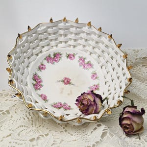 Könnte beinhalten: Weißer Keramikkorb-Schale mit geflochtenem Design und goldenen Verzierungen. Das Innere zeigt ein Blumenmuster mit rosa Rosen. Zwei getrocknete lila Rosen liegen in der Nähe der Schale, was auf ein Vintage-Thema hindeutet.