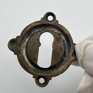 May include: A circular brass keyhole escutcheon plate with a weathered, dark patina. The hardware features a central vertical keyhole opening and three small mounting holes positioned around the outer edge for secure attachment to a door surface.