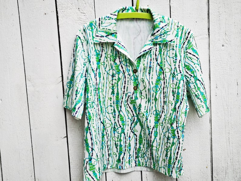 May include: A white short-sleeved shirt with a green and blue abstract pattern. The shirt has a collared neckline and a button closure.
