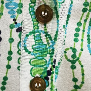 May include: Close-up of a white fabric with a green and blue pattern. Two gold buttons are visible on the fabric.