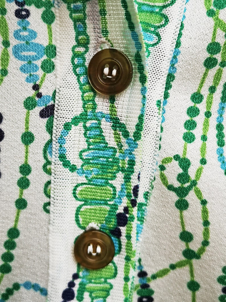 May include: Close-up of a white fabric with a green and blue pattern. Two gold buttons are visible on the fabric.
