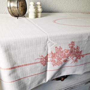 Könnte beinhalten: Eine weiße Tischdecke mit einem rosafarbenen Blumendesign und einem roten Rand. Die Tischdecke hat in der Mitte ein kleines ovales Design.