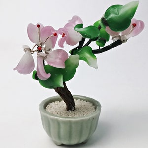 May include: A small glass bonsai tree with pink flowers and green leaves in a green ceramic pot. The tree is made of glass and has a brown wire trunk.