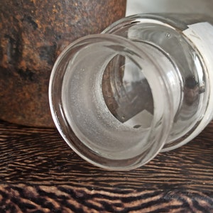 May include: A clear glass bottle with a frosted rim and a white label. The bottle is sitting on a dark wooden surface.