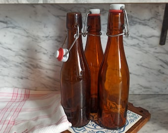 May include: Three brown glass bottles with swing top lids. The bottles are on a wooden surface with a blue and white patterned tile.