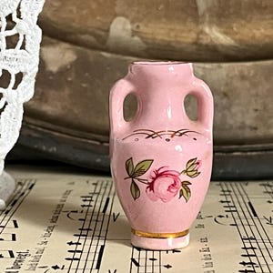 Könnte beinhalten: Kleine, rosa Keramikvase mit zwei Henkeln. Die Vase ist mit einer rosa Rose und grünen Blättern sowie goldenen Akzenten verziert. Die Vase steht auf einem Notenblatt.