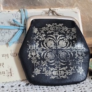 Könnte beinhalten: Eine schwarze Ledermünzbörse mit einem silbernen Verschluss und einem silbernen Blumendesign.