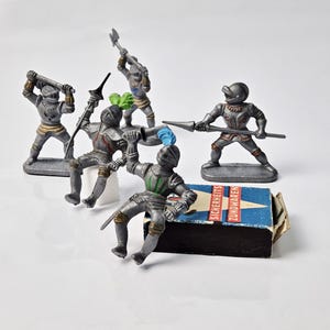 May include: A set of five silver toy knights with various weapons, including a spear, sword, and axe. The knights are standing on a small, blue and white box with the text "Sicherheits Zündwaren" printed on it.