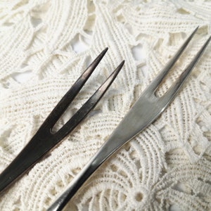 May include: Two silver forks with two tines each, one fork is polished and the other is tarnished, lying on a white crocheted surface.