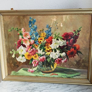 Op de afbeelding: Een ingelijste vintage print van een stilleven met een boeket kleurrijke bloemen in een heldere glazen vaas. De bloemen omvatten witte lelies, rode en roze rozen, gele en oranje madeliefjes en blauwe riddersporen. De bloemen zijn gerangschikt op een groene doek.