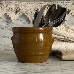 Könnte beinhalten: Ein brauner Keramik-Utensilienhalter mit glänzendem Finish, der Besteck enthält. Der Halter hat zwei kleine Griffe und ist mit Löffeln, Gabeln und einem Messer gefüllt. Ein Glas und gefaltete Handtücher befinden sich im Hintergrund.