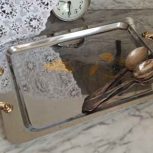 Könnte beinhalten: Ein silbernes Serviertablett mit goldenen Griffen und einem geätzten Blumenmuster. Zwei silberne Löffel liegen auf dem Tablett.